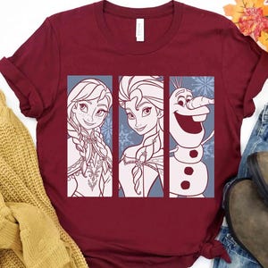 May include: A maroon short-sleeved t-shirt featuring a graphic print of three vertical panels. The panels display line art illustrations of Anna, Elsa, and Olaf from the movie Frozen, set against a light blue background with snowflake patterns.