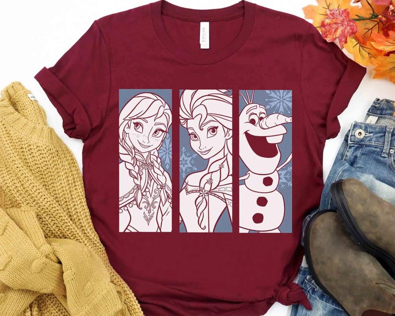 May include: A maroon short-sleeved t-shirt featuring a graphic print of three vertical panels. The panels display line art illustrations of Anna, Elsa, and Olaf from the movie Frozen, set against a light blue background with snowflake patterns.