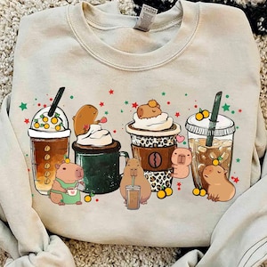 May include: Beige sweatshirt featuring a whimsical design of capybaras and coffee drinks. The capybaras are depicted with various coffee beverages, including iced coffee, lattes, and cappuccinos. The design includes orange and yellow accents, with red and green star details.