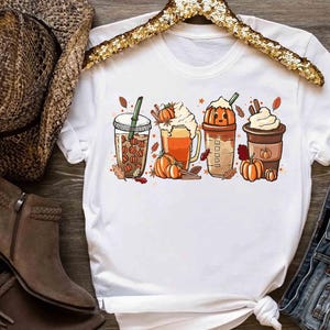 May include: A white short-sleeve t-shirt featuring a graphic print of four autumn-themed beverages. The design includes iced coffee with pumpkin shapes, a pumpkin-topped latte, a pumpkin-shaped cup, and a hot cocoa with whipped cream and pumpkins.