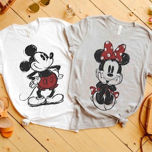 May include: Two t-shirts featuring cartoon characters. One is white with a black and red Mickey Mouse design. The other is gray with a Minnie Mouse design, including a red polka dot bow. Both shirts are short-sleeved.
