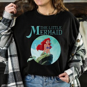 Pode incluir: T-shirt preto de gola redonda com um gr&aacute;fico de Ariel de "A Pequena Sereia". O design inclui o texto "The Little Mermaid" em verde-azulado e uma imagem circular de Ariel com cabelo ruivo, um suti&atilde; de concha roxa e uma cauda verde.