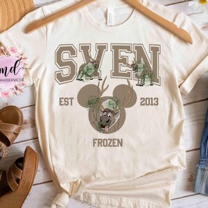 May include: Cream-colored t-shirt with a graphic design featuring the name "SVEN" in brown block letters above cartoon reindeer illustrations. Below, a cartoon reindeer face with antlers and the word "FROZEN". The shirt also has the text "EST 2013".