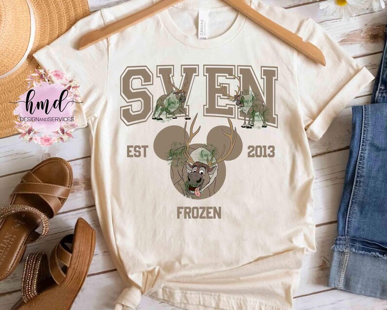 May include: Cream-colored t-shirt with a graphic design featuring the name "SVEN" in brown block letters above cartoon reindeer illustrations. Below, a cartoon reindeer face with antlers and the word "FROZEN". The shirt also has the text "EST 2013".