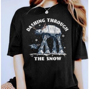 Może przedstawiać: Czarny t-shirt z białym nadrukiem AT-AT z rogami renifera, płatkami śniegu i napisem "Dashing Through The Snow". Projekt ma świąteczny motyw.
