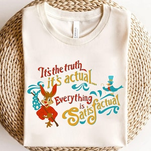 May include: Off-white t-shirt with a cartoon design of a rabbit in a red outfit and a blue bird wearing a top hat. The shirt displays the text "It's the truth, it's actual, everything is satisfactual."