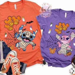 Op de afbeelding: Twee Halloween-t-shirts. E&eacute;n is oranje met een mummie-achtig Stitch-personage dat Mickey Mouse pompoenballonnen vasthoudt. De andere is paars met een roze Angel-personage dat Mickey Mouse pompoenballonnen vasthoudt. Beide shirts hebben spinnenwebben, geesten en vleermuizen.