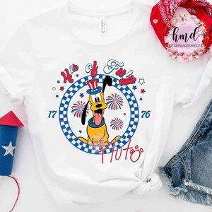 May include: White t-shirt with a cartoon Pluto design, celebrating the 4th of July. The design includes the text "4th of Fun", fireworks, stars, and the numbers "77" and "76". The colour scheme is red, white, and blue.