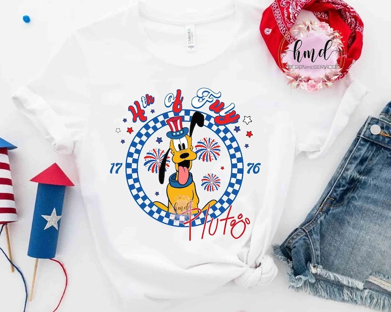 May include: White t-shirt with a cartoon Pluto design, celebrating the 4th of July. The design includes the text "4th of Fun", fireworks, stars, and the numbers "77" and "76". The colour scheme is red, white, and blue.