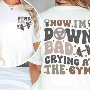 May include: Off-white t-shirt with the words "DOWN BAD" and a barbell graphic on the front. The back of the shirt displays the phrase "NOW I'M DOWN BAD CRYING AT THE GYM" in a retro font, with dumbbell and kettlebell graphics.