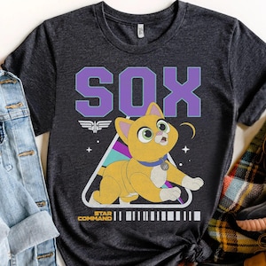 Peut inclure: T-shirt gris foncé avec le mot "SOX" en lettres violettes au-dessus d'une illustration de chat de dessin animé. Le chat est jaune avec des yeux verts et un collier bleu, sur un triangle aux accents arc-en-ciel. Le texte "STAR COMMAND" est en dessous.