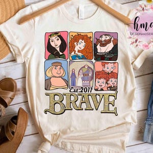 May include: Cream-colored t-shirt featuring a colorful graphic of characters from the animated film "Brave." The design includes six character portraits and the word "BRAVE" in a decorative font, with "Est. 2011" below.