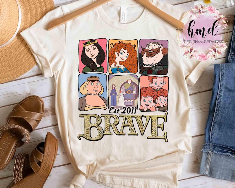 May include: Cream-colored t-shirt featuring a colorful graphic of characters from the animated film "Brave." The design includes six character portraits and the word "BRAVE" in a decorative font, with "Est. 2011" below.