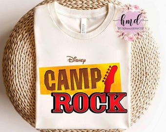 May include: Cream-colored t-shirt featuring the Disney Camp Rock logo. The design includes the words "CAMP ROCK" in brown and red, with a guitar neck graphic. The shirt is displayed on a woven surface.