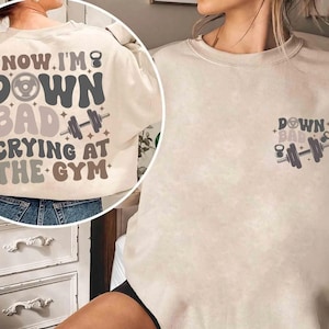 May include: A cream-coloured sweatshirt with the text "NOW I'M DOWN BAD CRYING AT THE GYM" in a retro font. The back of the shirt features a large graphic with a dumbbell, kettlebell, and stars. A smaller version of the graphic is on the front.