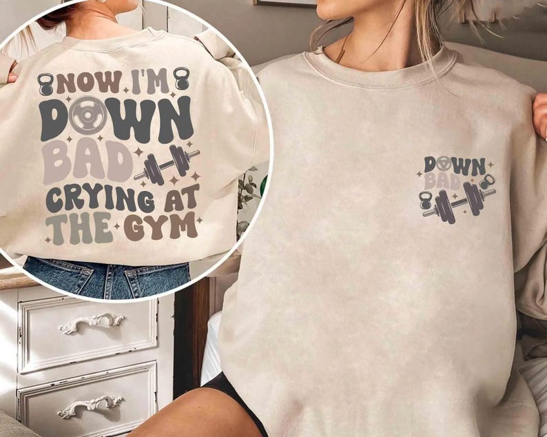 May include: A cream-coloured sweatshirt with the text "NOW I'M DOWN BAD CRYING AT THE GYM" in a retro font. The back of the shirt features a large graphic with a dumbbell, kettlebell, and stars. A smaller version of the graphic is on the front.