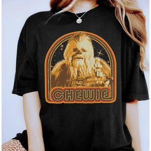 May include: Black t-shirt with a retro graphic of Chewbacca and the word "CHEWIE" in orange. The design features a brown and orange arch with stars, creating a vintage look. The t-shirt is a classic fit.