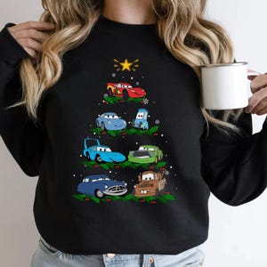 May include: Black sweatshirt featuring a Christmas tree design made of cartoon cars. The cars are various colors, including red, blue, and green, with a yellow star at the top. The design includes snowflakes and holly.