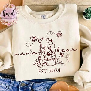 May include: Cream-colored sweatshirt featuring a Winnie the Pooh design with the words "mama bear" and "Hunny". The design includes bees and the year "EST. 2024". The sweatshirt is made of a soft material.