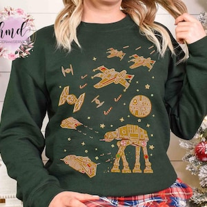 Puede incluir: Sudadera verde oscuro con un diseño temático de Star Wars. El diseño presenta ilustraciones doradas de naves espaciales, la Estrella de la Muerte y un caminante AT-AT, con detalles de bastones de caramelo. Es un artículo festivo de vacaciones.