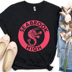 Puede incluir: Camiseta negra con un gráfico rosa desgastado de un camarón dentro de un círculo, con las palabras "SEABROOK HIGH". La imagen también muestra bailarinas rosas y vaqueros azules.