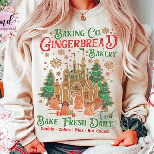 Pode incluir: Camisola bege com um design festivo de padaria de gengibre. O gráfico inclui um castelo de gengibre, árvores de Natal, bonecos de gengibre e o texto "Baking Co. Gingerbread Bakery. Bake Fresh Daily."