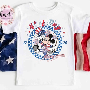 May include: White t-shirt with a patriotic Mickey Mouse design. The graphic includes the text "4th of July" and "1776" in red and blue, with fireworks and stars. Mickey Mouse is depicted in patriotic clothing, celebrating Independence Day.