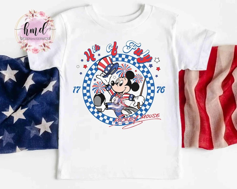 May include: White t-shirt with a patriotic Mickey Mouse design. The graphic includes the text "4th of July" and "1776" in red and blue, with fireworks and stars. Mickey Mouse is depicted in patriotic clothing, celebrating Independence Day.
