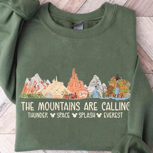 May include: Green sweatshirt with a graphic of colorful mountain illustrations and the text "THE MOUNTAINS ARE CALLING" with the words THUNDER, SPACE, SPLASH, and EVEREST below. The sweatshirt is made of a soft material.