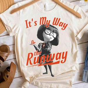 May include: Cream-colored t-shirt featuring a cartoon character with large glasses and a black bob. The text "It's My Way Or the Runway" is printed in red. The shirt is hanging on a wooden hanger.