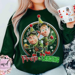 May include: A dark green sweatshirt featuring a circular graphic with two cartoon elves in green outfits, goggles, and hats. The graphic includes the text "Prep & Landing" in red and green. A white mug with a red handle and a Nutcracker design is also visible.