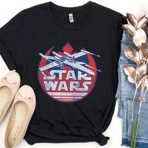 May include: Black t-shirt featuring a distressed graphic of a Star Wars X-wing fighter inside a red, white, and blue circle. The words "STAR WARS" are printed below the fighter. The shirt is paired with pink ballet flats and blue jeans.