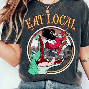 May include: Dark gray t-shirt featuring a cartoon of Captain Hook running from a crocodile, with the words "EAT LOCAL" in a circular design. The design is in shades of red, green, and yellow.