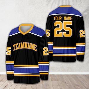Pu&ograve; includere: Maglia da hockey nera con strisce blu e dorate. La maglia ha il testo "TEAMNAME" sul davanti e il numero "25" sul retro con il testo "YOUR NAME" sopra.