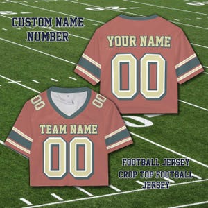 Puede incluir: Dos camisetas de f&uacute;tbol rojas cortas con el texto "TEAM NAME" y "YOUR NAME" sobre el n&uacute;mero "00". Las camisetas tienen rayas verdes y beige en las mangas. Tambi&eacute;n es visible el texto "CUSTOM NAME NUMBER" y "FOOTBALL JERSEY CROP TOP FOOTBALL JERSEY".