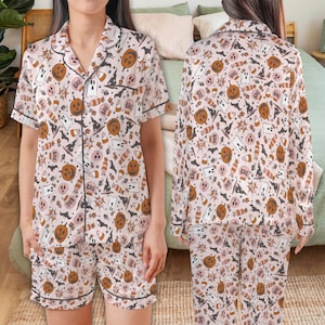 Puede incluir: Conjunto de pijama rosa con temática de Halloween con un patrón repetido de calabazas, fantasmas, caramelos y sombreros de bruja. El conjunto incluye una camisa de manga corta con botones y pantalones cortos, ambos con ribete negro.