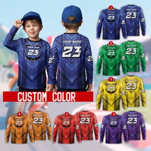 May include: A young child wearing a blue long-sleeve shirt with the number 23 and the text "YOUR TEXT" on the front. The shirt has a geometric design. The image also shows several other shirts in different colors, including green, yellow, red, and purple.