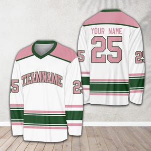 Pu&ograve; includere: Una maglia da hockey bianca con strisce rosa e verdi. La maglia ha il numero "25" sul retro e il testo "YOUR NAME" sopra. La parte anteriore della maglia ha il testo "TEAMNAME" in lettere rosa.