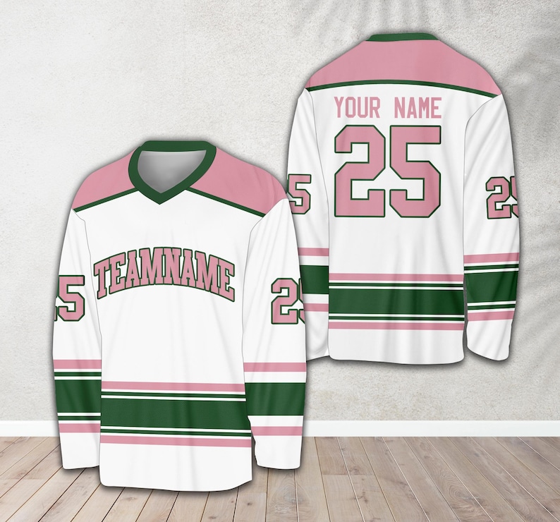 Pu&ograve; includere: Una maglia da hockey bianca con strisce rosa e verdi. La maglia ha il numero "25" sul retro e il testo "YOUR NAME" sopra. La parte anteriore della maglia ha il testo "TEAMNAME" in lettere rosa.