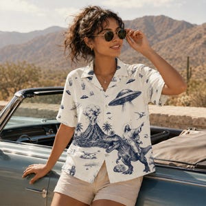 May include: Off-white button-down shirt with a whimsical blue illustration featuring a volcano, UFOs, and a figure riding a dinosaur. The shirt has short sleeves and a classic collar. The shirt is paired with beige shorts.