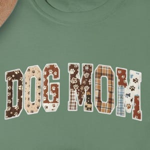 Op de afbeelding: Een lichtgroen t-shirt met de tekst "DOG MOM" in een patchwork ontwerp. De letters hebben pootafdrukken en ruitpatronen in bruin, beige en blauw. De tekst staat in het midden van het shirt.