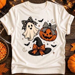 May include: A cream-colored short-sleeve t-shirt featuring a graphic print of a white ghost with a black polka-dot bow, an orange jack-o'-lantern with a black and white striped bow, and a black witch hat with an orange polka-dot bow.
