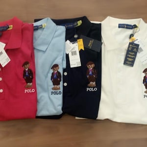 May include: Four polo shirts in red, light blue, navy, and white. Each shirt features a bear graphic and the word "POLO" embroidered on the front. The shirts have a classic collar and button placket.
