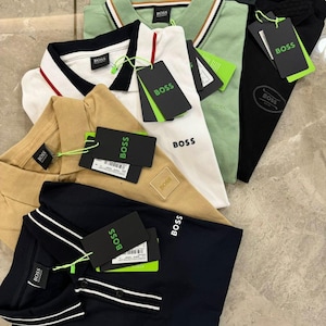 Puede incluir: Una colección de polos en varios colores, incluyendo negro, azul marino, blanco, beige y verde. Cada camisa tiene un cuello y el logo "BOSS". Algunas camisas tienen detalles a rayas en el cuello y las mangas, y todas tienen etiquetas adjuntas.