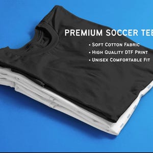 K&ouml;nnte beinhalten: Ein Stapel aus zwei T-Shirts, eines schwarz und eines wei&szlig;, vor blauem Hintergrund. Das schwarze Shirt oben zeigt "PREMIUM SOCCER TEE" und Details zu Stoff und Passform. Die Shirts sind sauber gefaltet.