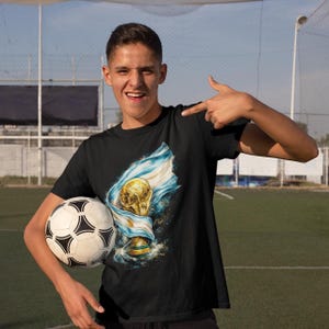 K&ouml;nnte beinhalten: Schwarzes T-Shirt mit einer Grafik einer goldenen Troph&auml;e und einer blau-wei&szlig;en Flagge, mit einem Fu&szlig;ball im Vordergrund. Das Design feiert eine Fu&szlig;ballmeisterschaft.