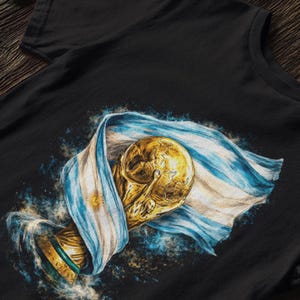 K&ouml;nnte beinhalten: Schwarzes T-Shirt mit einer detaillierten Illustration einer goldenen Troph&auml;e, die in die argentinische Flagge geh&uuml;llt ist. Das Design umfasst blaue und wei&szlig;e Streifen, ein Sonnenemblem und einen Spritzeffekt.