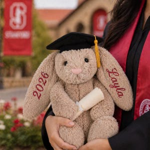 Puede incluir: Un peluche de conejo beige con un gorro de graduación negro con borla dorada. El conejo sostiene un diploma enrollado y tiene bordados "2026" y "Layla". Un regalo de graduación.