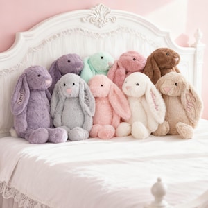 Può includere: Una collezione di coniglietti di peluche in vari colori pastello, tra cui lavanda, grigio, verde menta, rosa, bianco e marrone. I coniglietti sono seduti su un letto bianco con una testiera decorativa. Alcuni coniglietti hanno nomi ricamati sulle orecchie.