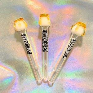 Könnte beinhalten: Drei transparente Glas-Raucherpfeifen mit weißen und gelben Akzenten. Jede Pfeife trägt den schwarzen Aufdruck "GOOVYGLASS". Die Pfeifen sind auf einem holografischen, irisierenden Hintergrund angeordnet.